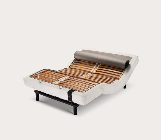 An adjustable bed base with wooden slats visible, covered by a beige fabric. The bed base is designed to be adjustable for different firmness levels and includes a wireless remote control.