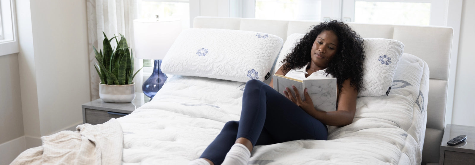 Woman relaxes in a seated position on a PranaSleep mattress with the adjustable base elevated, reading comfortably in bed and showcasing customizable head support for personalized comfort and relaxation.